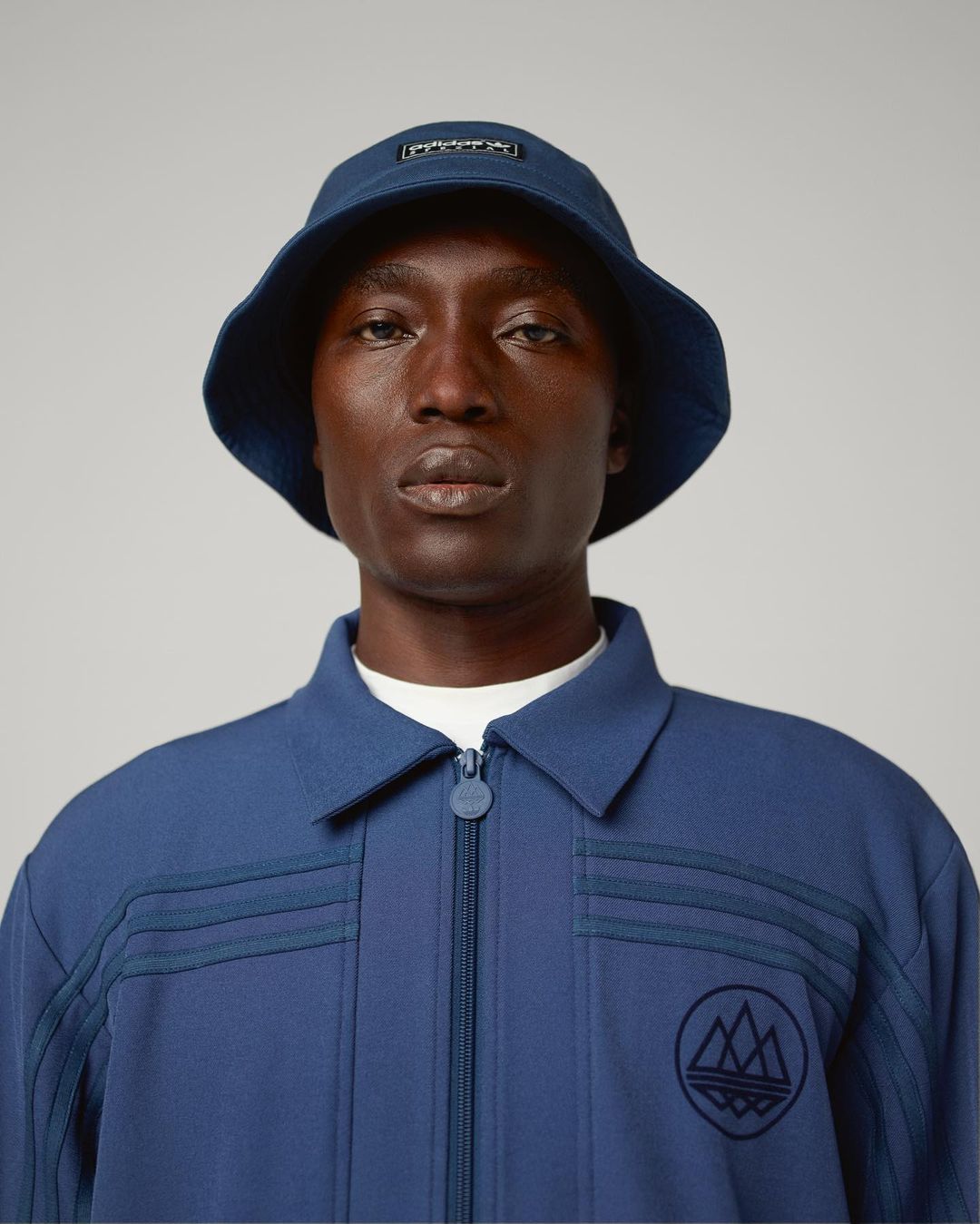 Dark-skinned adult wears a blue bucket hat and zip-up jacket with embroidered logo. Indoor shot against a gray backdrop.