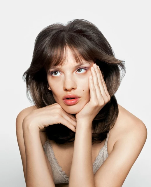 Headshot of a young woman with brown hair, lace top, and pink eyeshadow, resting her face on her hands against a plain white background.