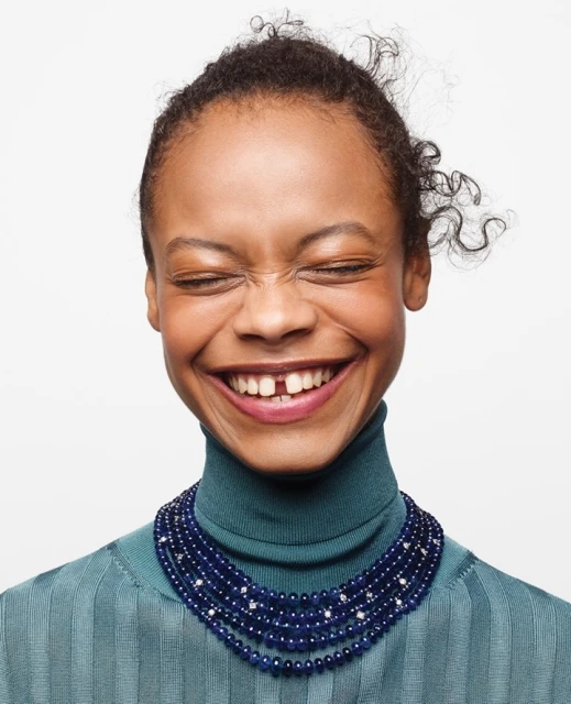 Close-up studio shot of a smiling African-American woman wearing a teal turtleneck and a layered sapphire bead necklace against a white background.