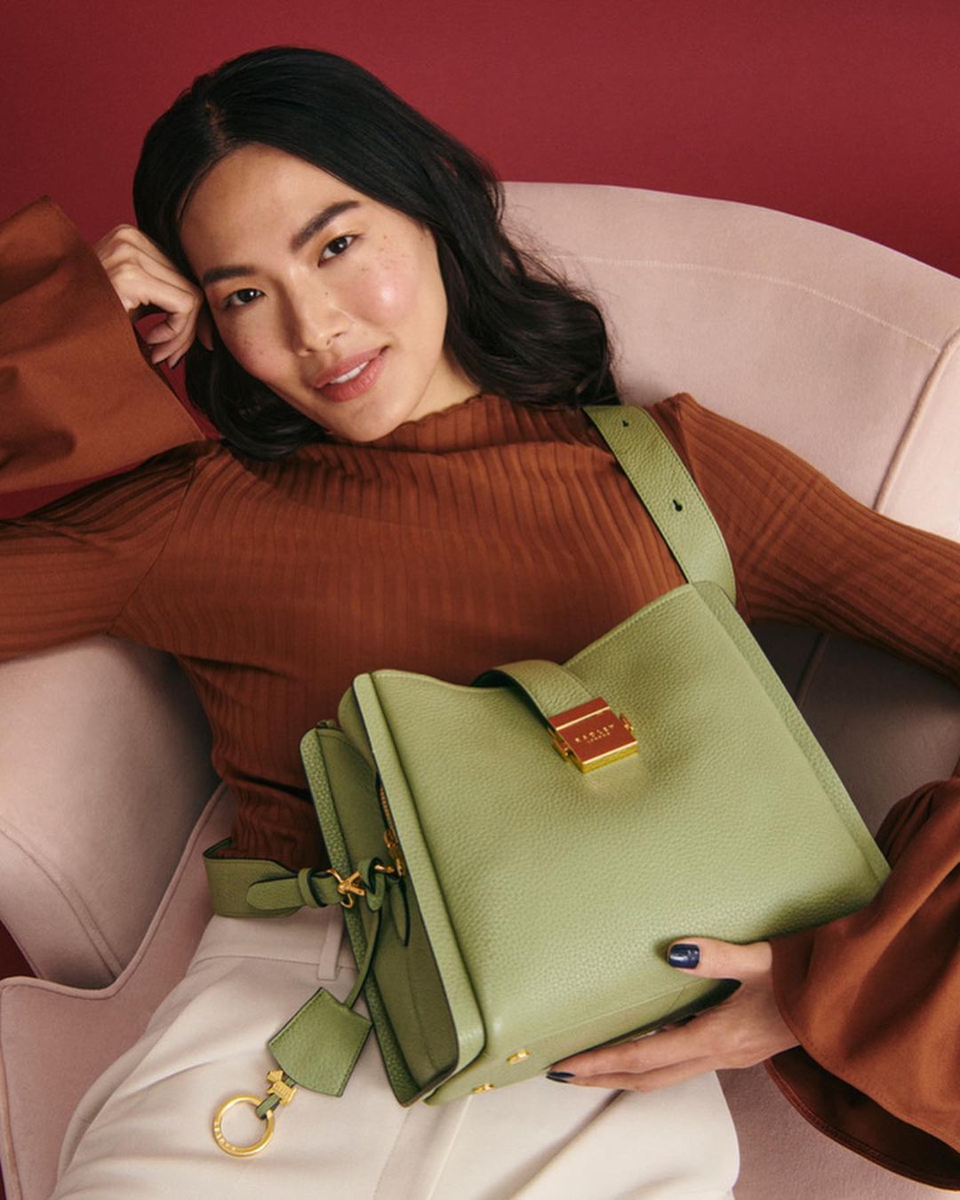 Adult woman reclines in chair holding a green leather handbag with gold hardware and a brown top and white pants in a studio.