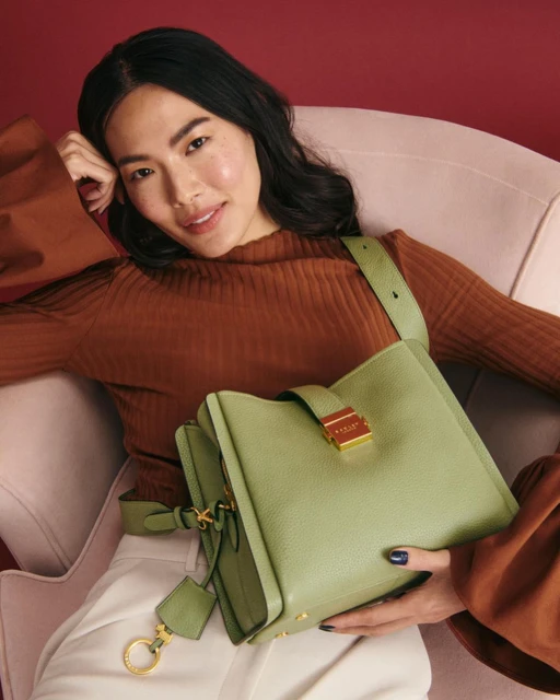 Adult woman reclines in chair holding a green leather handbag with gold hardware and a brown top and white pants in a studio.