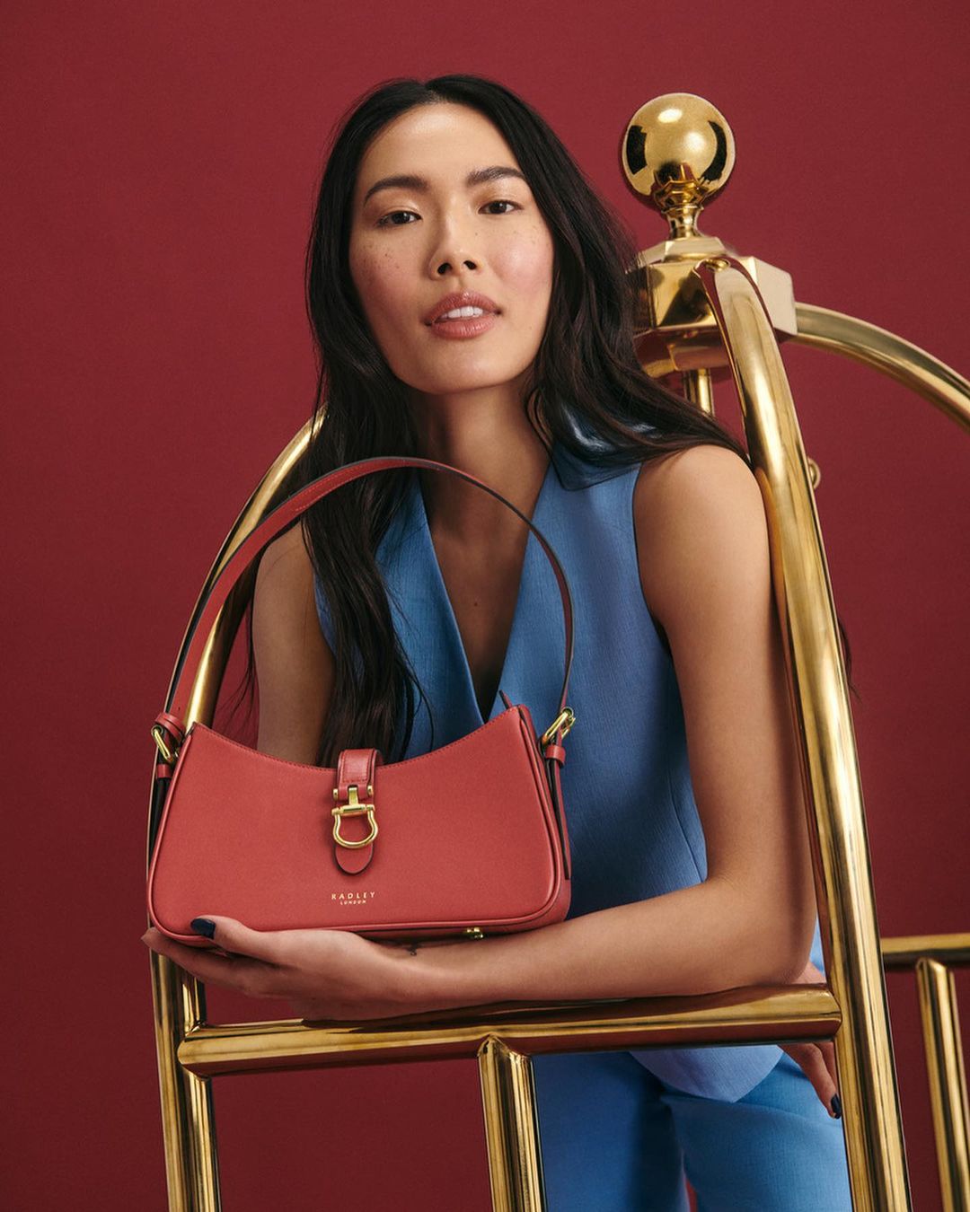Woman with an Asian appearance holds a coral-colored Radley purse next to a gold-toned luggage cart against a red backdrop.