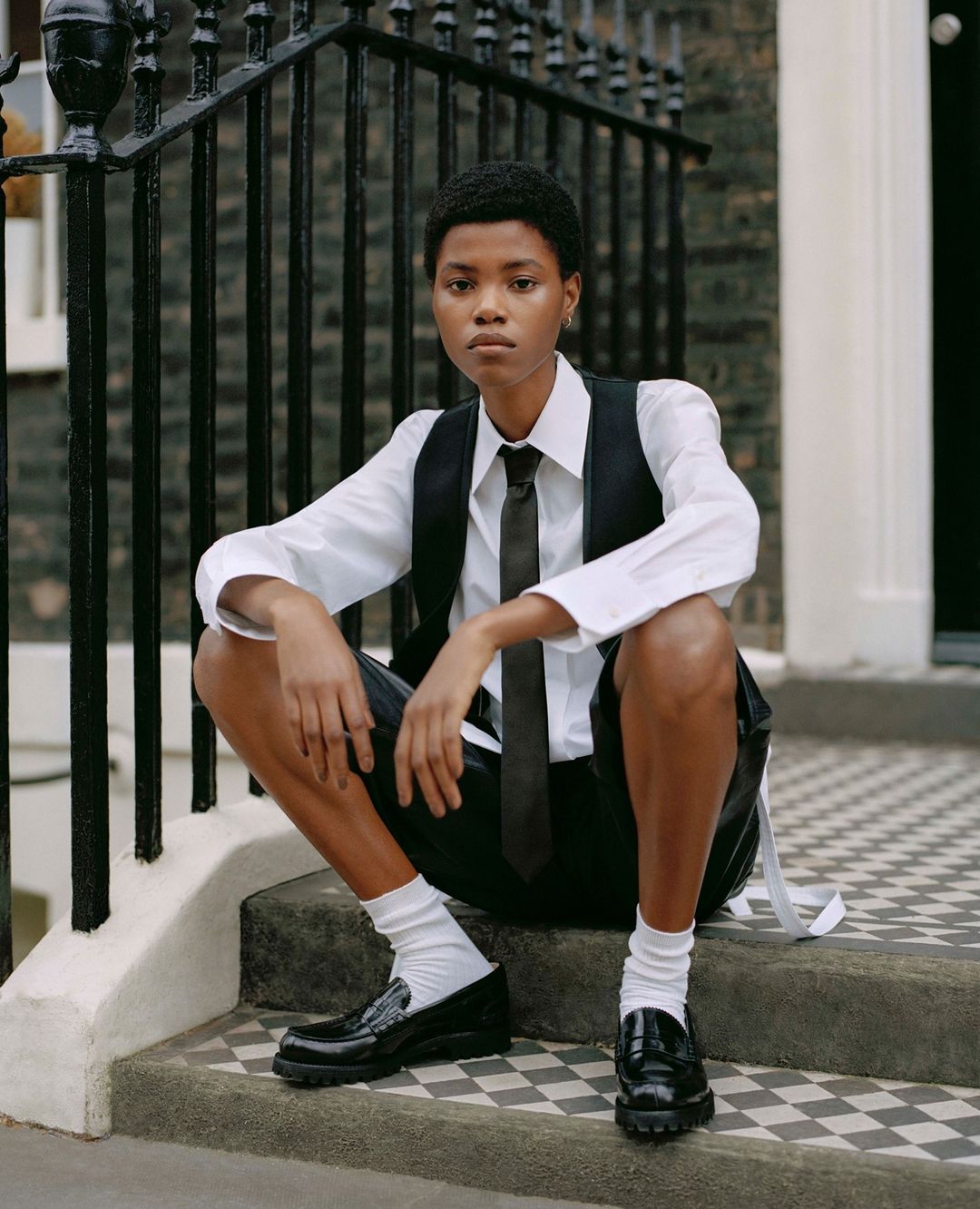 Woman in white shirt, vest, tie, and shorts sits on outdoor steps with black loafers and white socks, posing for a fashion shot.