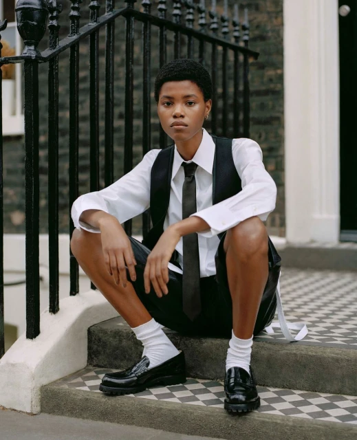 Woman in white shirt, vest, tie, and shorts sits on outdoor steps with black loafers and white socks, posing for a fashion shot.