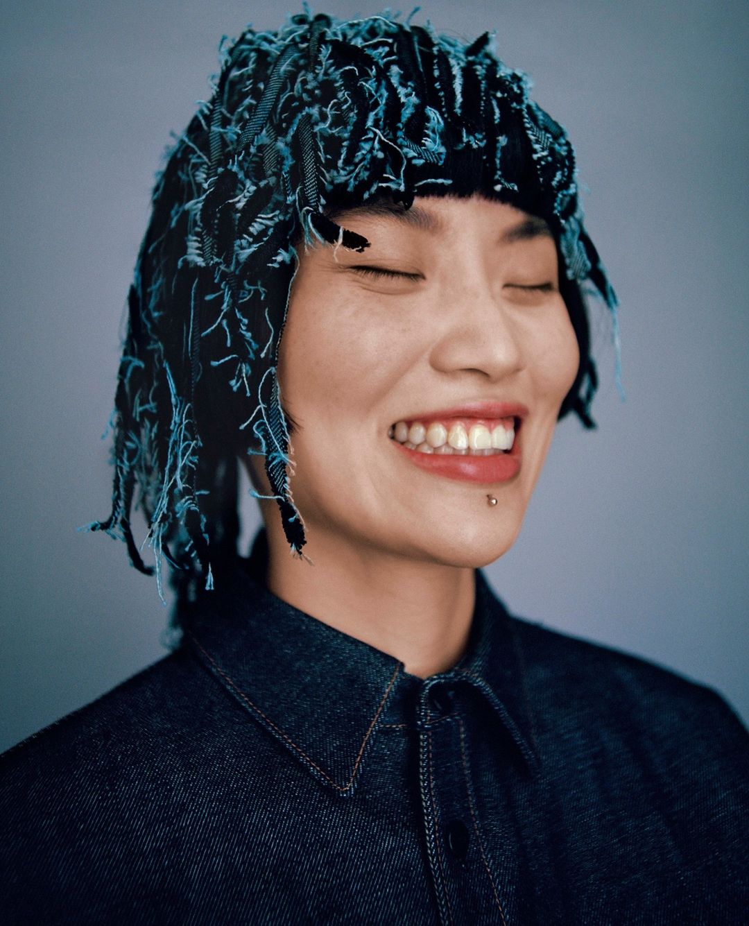 Portrait of a smiling Asian woman with closed eyes, wearing a denim shirt and a blue and black decorative headpiece indoors.
