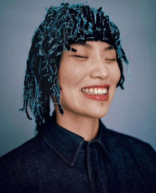 Portrait of a smiling Asian woman with closed eyes, wearing a denim shirt and a blue and black decorative headpiece indoors.