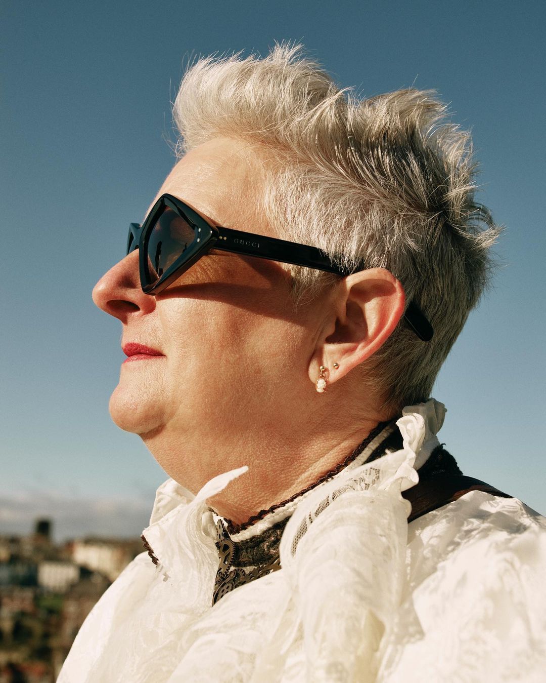 Side profile of a woman wearing black Gucci sunglasses, white ruffled top, and stud earrings, set against a clear blue sky.