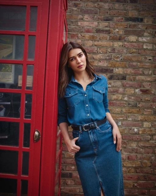 Woman in a denim shirt and skirt poses against a red telephone booth and brick wall, showcasing a blend of casual fashion and urban backdrop.