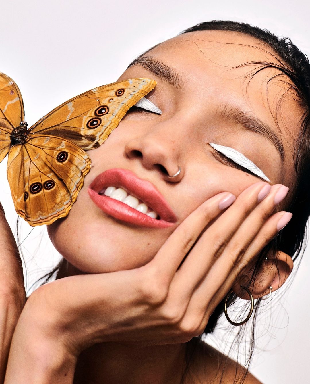 Close-up beauty shot of an adult woman with makeup, hand near her face and a butterfly over her eye against a bright white background.