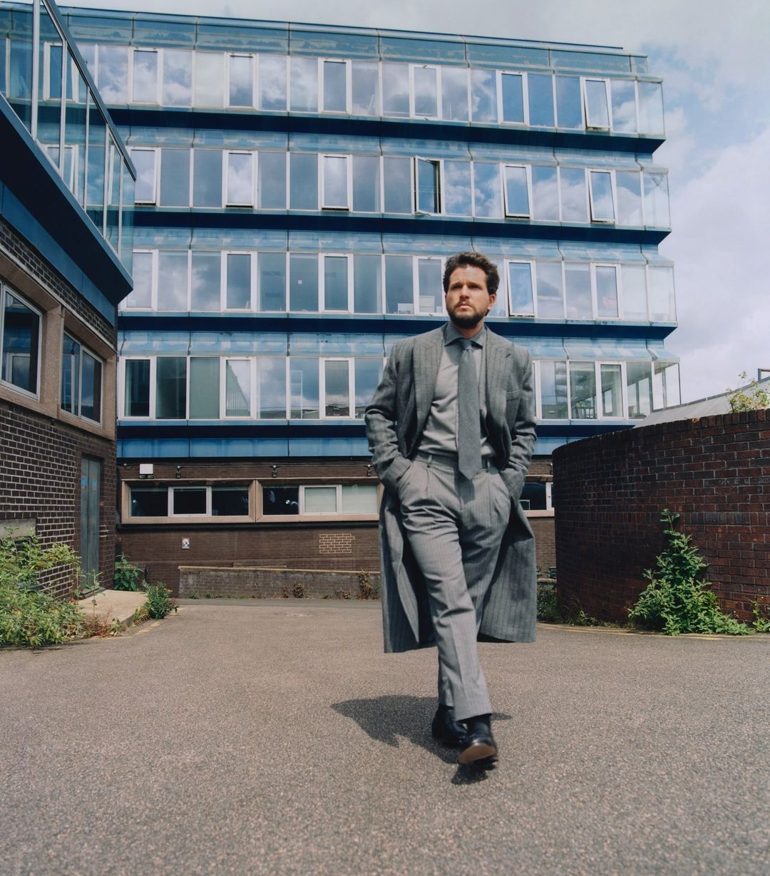 Adult male in a gray suit and long coat walking on a paved area in front of a modern building with many windows on a sunny day.