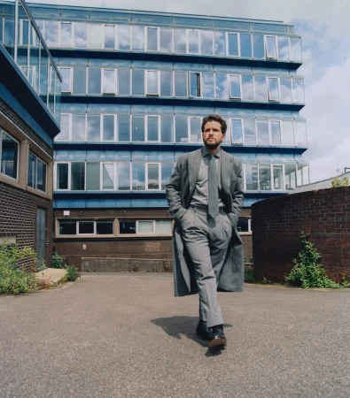 Adult male in a gray suit and long coat walking on a paved area in front of a modern building with many windows on a sunny day.