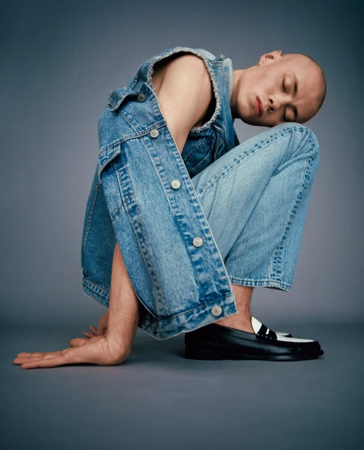 Bald adult wearing denim vest and jeans in a studio setting. The subject is kneeling with hands on the ground and eyes closed.