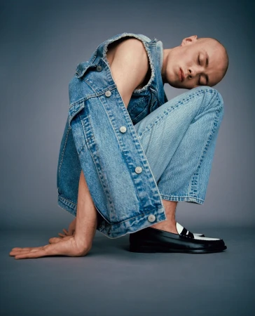 Bald adult wearing denim vest and jeans in a studio setting. The subject is kneeling with hands on the ground and eyes closed.