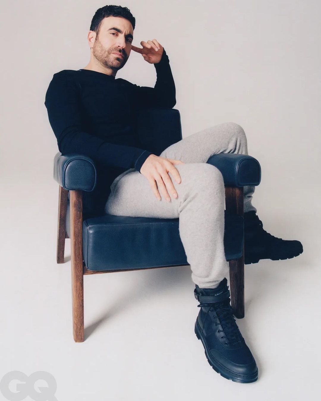 Portrait of adult male with dark hair and beard, wearing a black long-sleeve shirt and grey sweatpants, seated in a blue chair in studio.