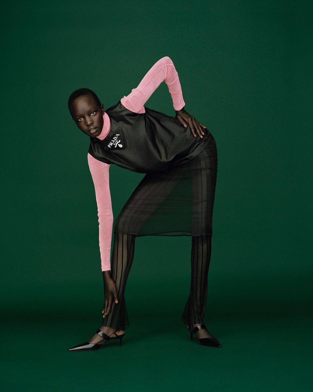Dark-skinned woman in a black Prada outfit with a pink long-sleeve undershirt poses on a deep green background in a studio fashion shot.