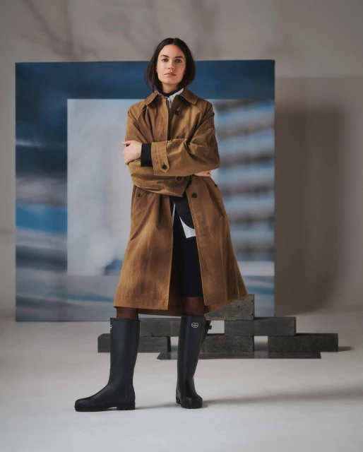 A stylish woman poses indoors wearing a tan trench coat, black boots, and tights against a backdrop with a gray and blue abstract pattern.
