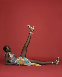 Black woman reclining on a red seamless background with one leg raised, wearing white strappy heels and a colorful sleeveless dress.
