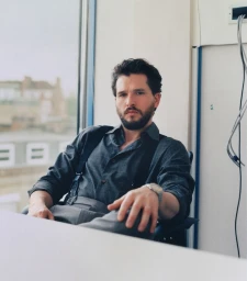 Kit Harington seated indoors wearing a dark grey shirt and suspenders with a window showing a street background
