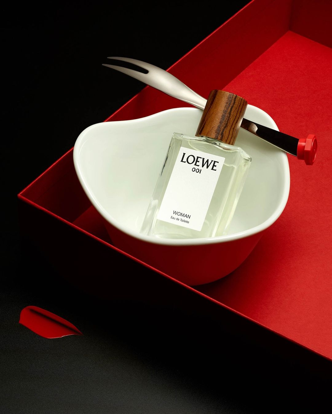 Still life shot of Loewe 001 Woman perfume in a white bowl, with a silver fork, resting inside a red box, contrasted by black backdrop.