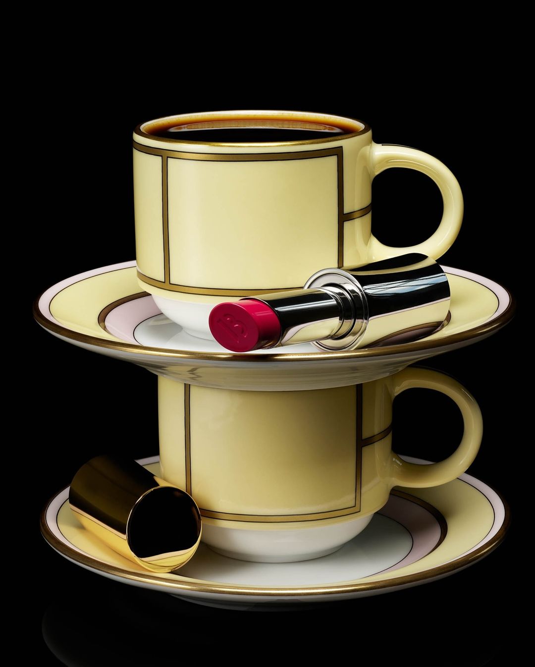 Elegant stack of two yellow teacups and saucers with lipstick, creating a beauty and beverage still life on a black backdrop.