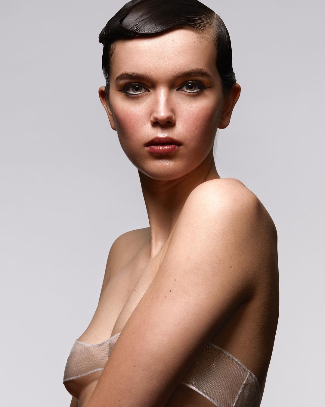 Striking beauty portrait of a young fair-skinned woman with dark, slicked-back hair and sheer white fabric accent on a light gray background.