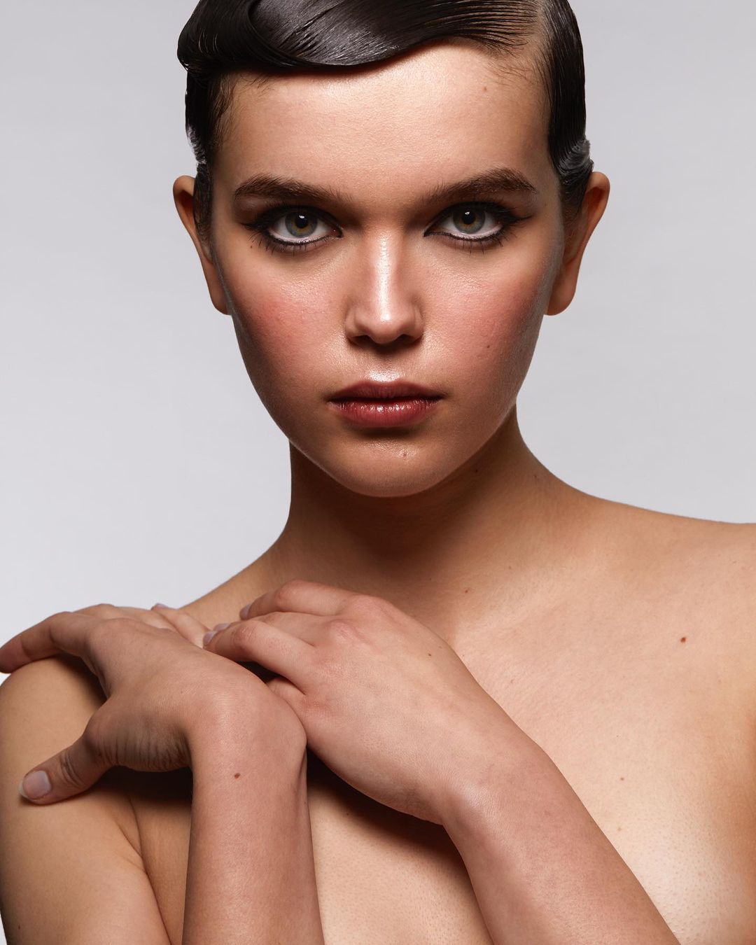 Close-up studio portrait of a woman with wet-look hair, dramatic makeup, and bare shoulders against a solid grey background.