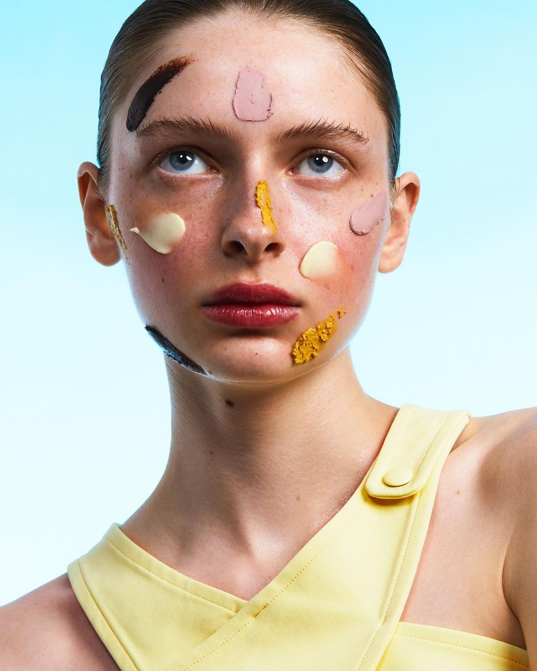Close-up of a fair-skinned woman with various cosmetic products applied to her face, shot against a gradient blue background.