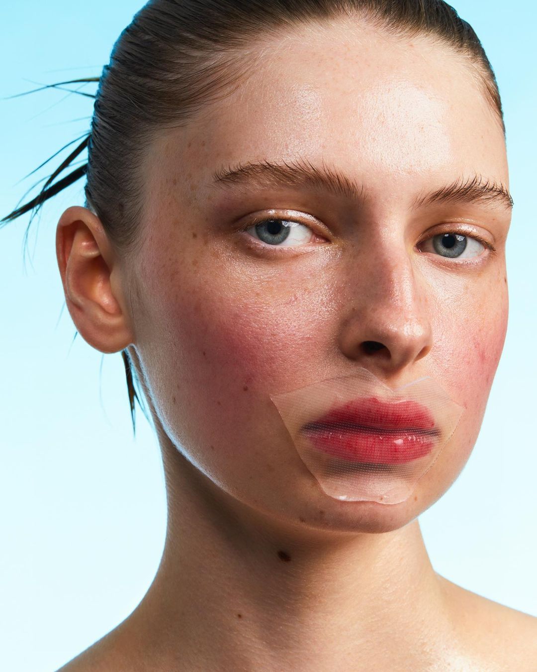 Close-up portrait of a fair-skinned young woman with blue eyes and red lips, wearing a transparent lip mask on a blue background.