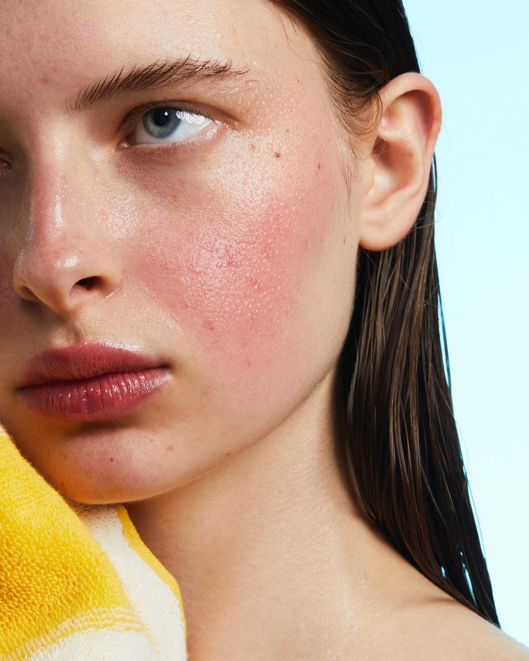 Close-up portrait of an adult woman with wet dark hair and pink cheeks, holding a yellow-and-white striped towel against a light blue background.