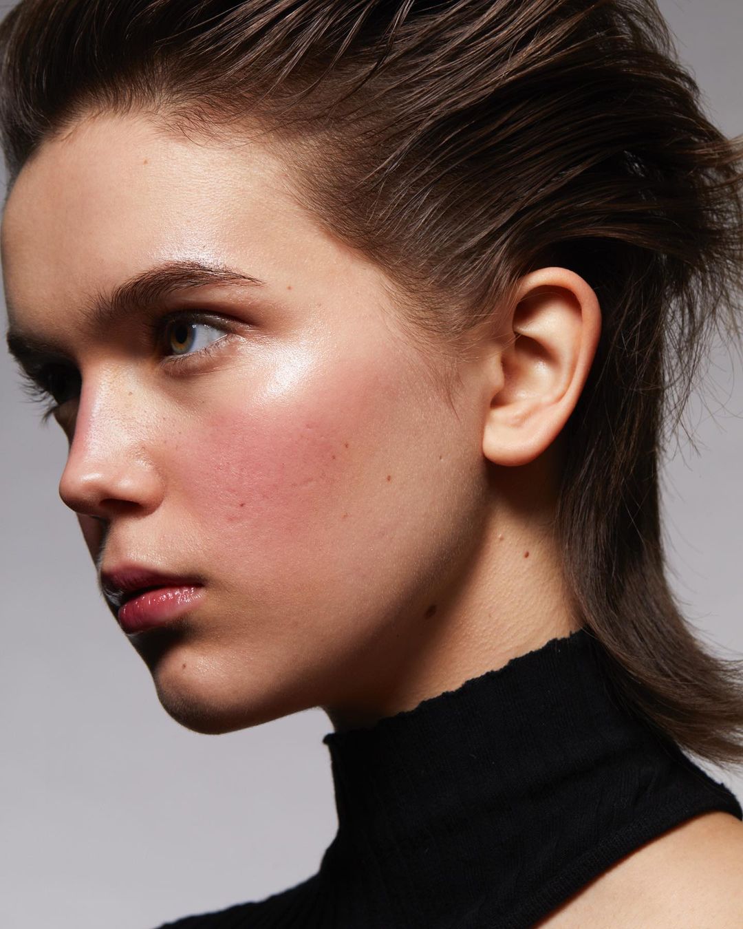 Close-up profile of a young woman with short, styled brown hair, wearing a black top against a neutral grey background. Beauty shot.