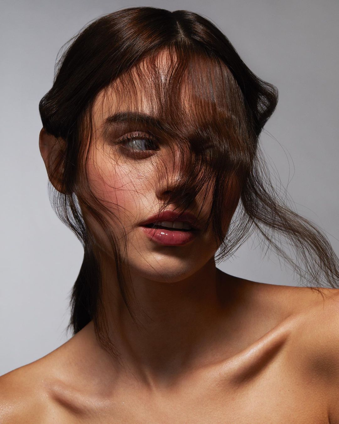 Close-up portrait of an adult female with dark hair covering part of her face, posing in a photo studio with neutral grey background.