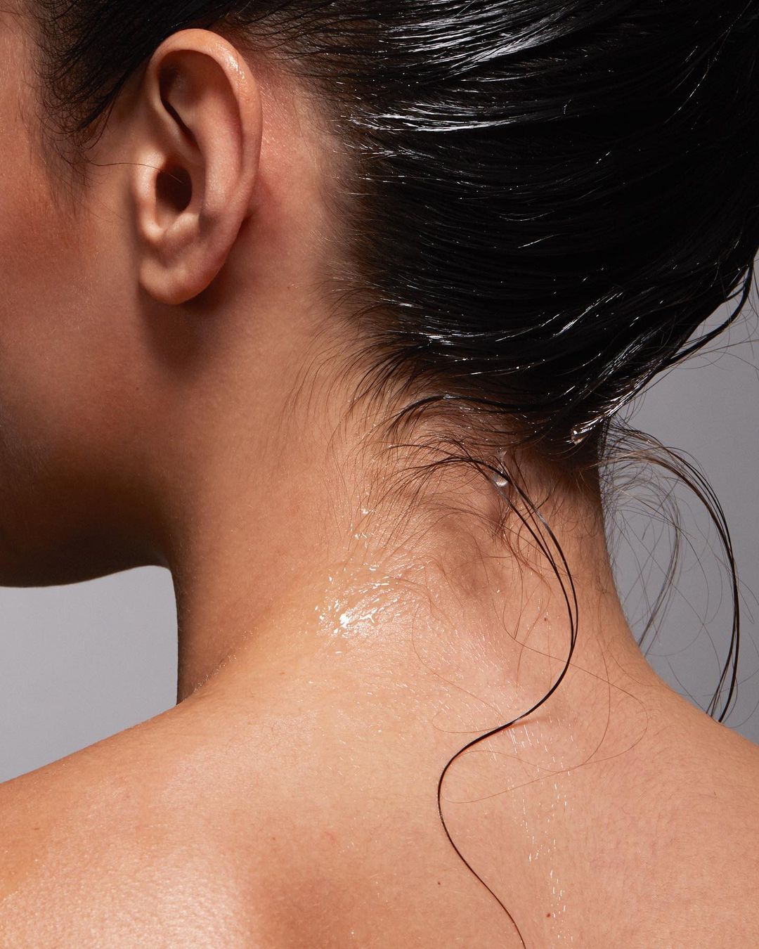 Close-up shot of a woman's neck and back, showcasing wet dark hair, skin texture, and glistening water droplets, set against a grey background.