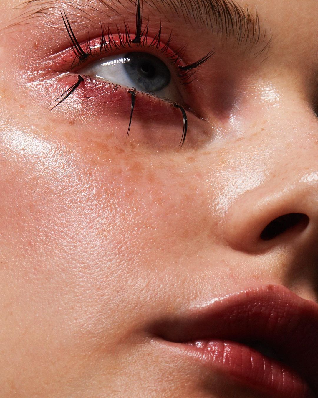 Close-up of a fair-skinned female face with freckles, red lipstick, and distinctive black eyelashes against a soft, neutral backdrop.