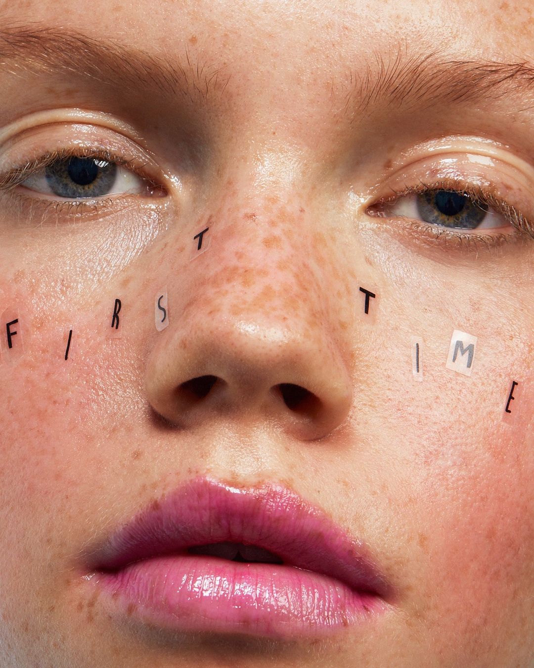 Close-up of a young woman's face with freckles, styled with letter stickers spelling 'first time' across her cheekbones and forehead.