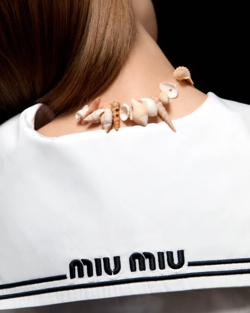 Woman's back view featuring a handmade seashell necklace, white top with the 'MIU MIU' logo, and a black background in soft studio lighting.