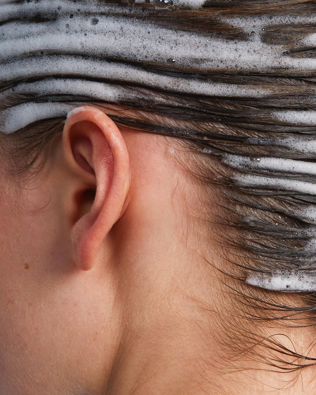Close-up shot of a fair-skinned person with brown hair covered in white shampoo suds, highlighting the ear and side of the face.
