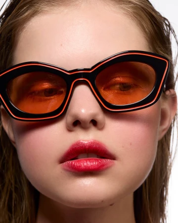Close-up portrait of a model wearing black geometric sunglasses with orange-tinted lenses and red lipstick against a plain white background.