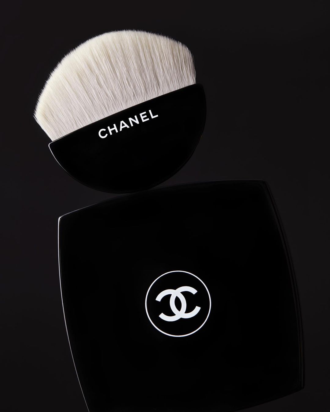 Chanel powder brush and compact case on a black surface. Beauty and cosmetics product photography with stark lighting.