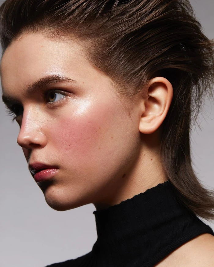 Close-up profile portrait of a woman with slicked-back brown hair and flushed pink cheeks wearing a black top against a grey background.