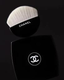 Close-up product shot of a Chanel makeup brush resting on a black compact case against a dark background.