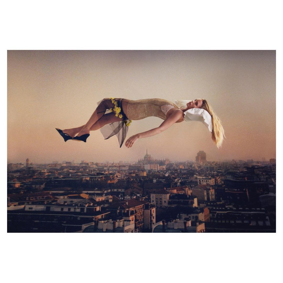 Woman in dress and heels floats above cityscape with pillow. Outdoor scene features aerial perspective, warm light, and urban architecture.
