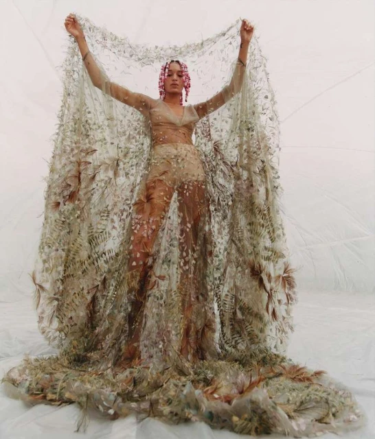 A woman stands in a light studio wearing a decorative dress made of foliage with a leaf-covered veil held up above her head.