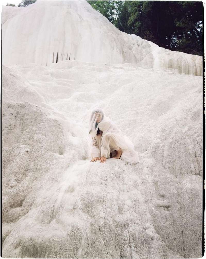 A woman in white clothing is sitting on a white mineral formation, with a forest visible in the background, in an eye-level shot.