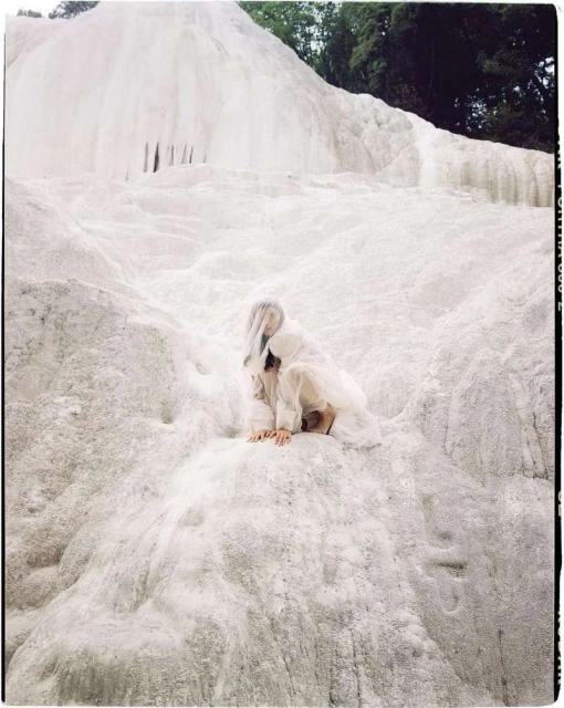 A woman in white clothing is sitting on a white mineral formation, with a forest visible in the background, in an eye-level shot.