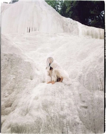 A woman in white clothing is sitting on a white mineral formation, with a forest visible in the background, in an eye-level shot.