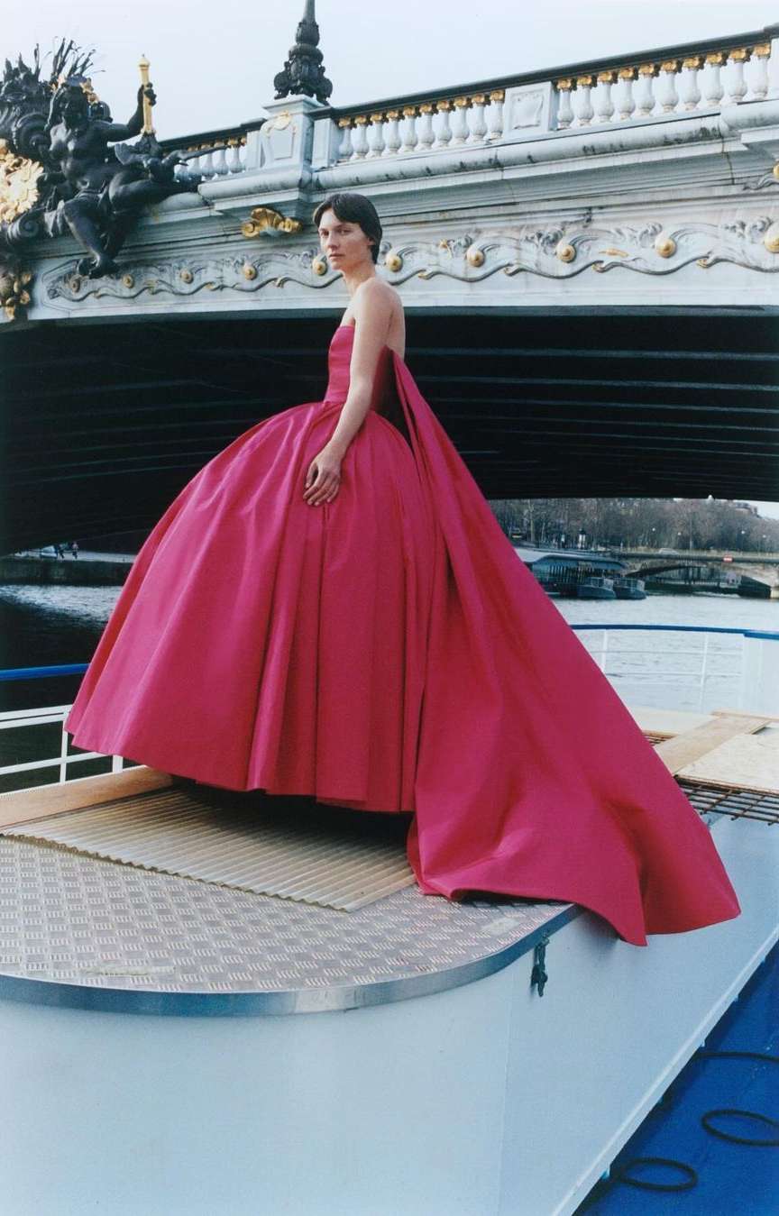 Woman poses in a pink ballgown on a boat with a bridge and river as a backdrop, creating a stunning fashion moment.
