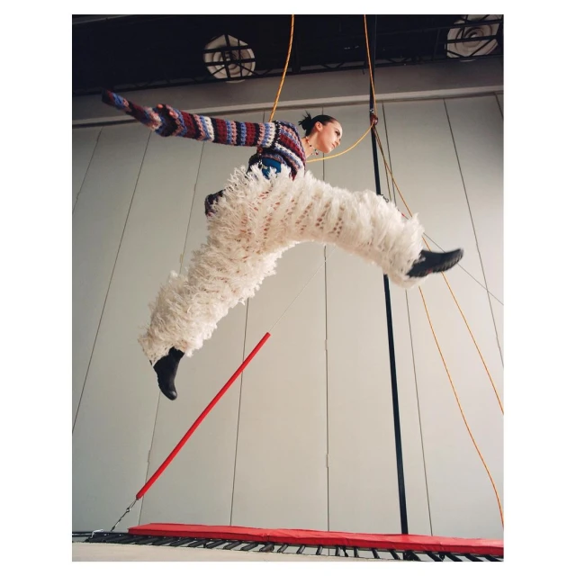 Woman in motion wearing a colorful knit sweater and fluffy white pants jumping on a red and black trampoline in an indoor space.