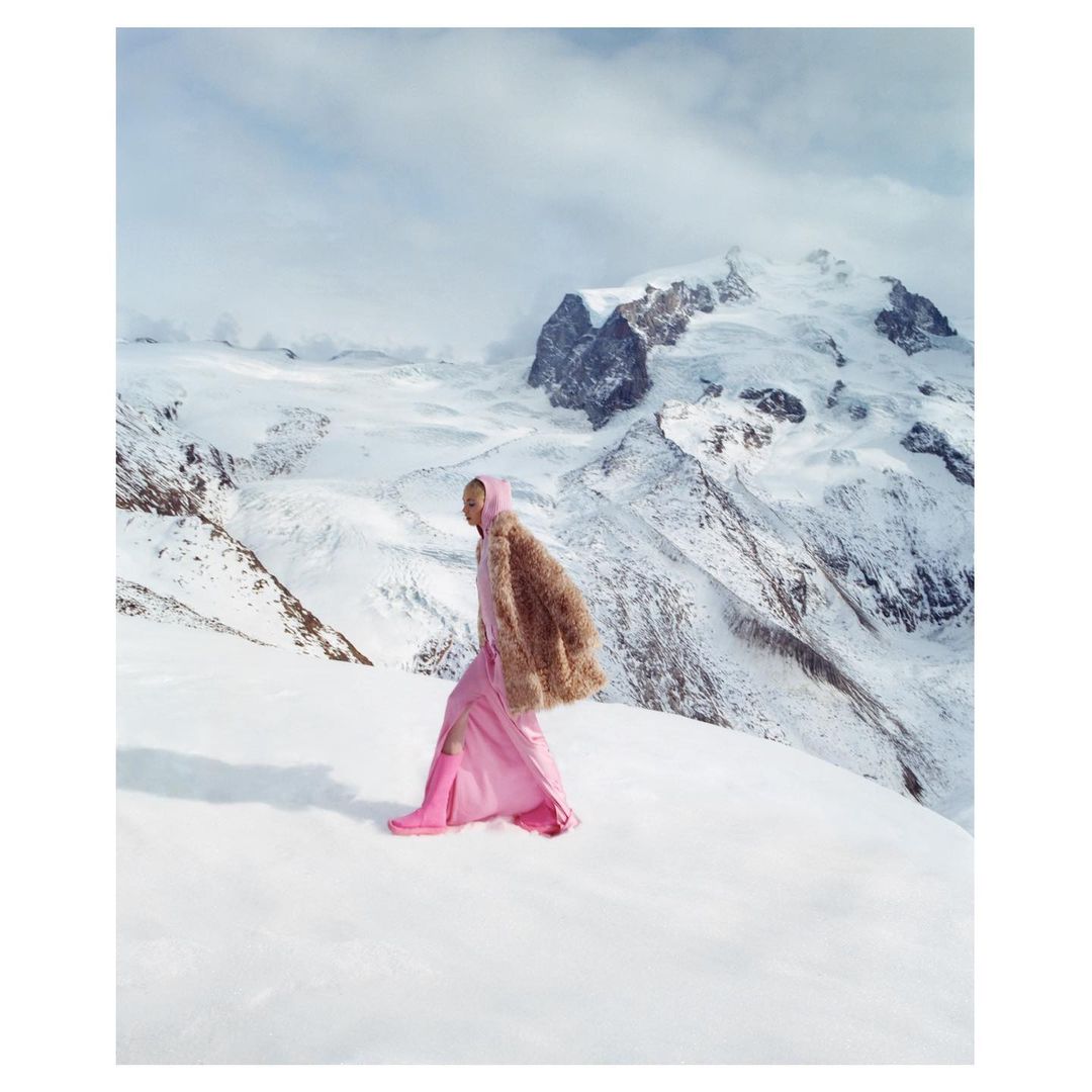 Fashionable woman in a pink dress and fur coat walks on snow-covered ground with snow-capped mountains and a bright sky in the background.
