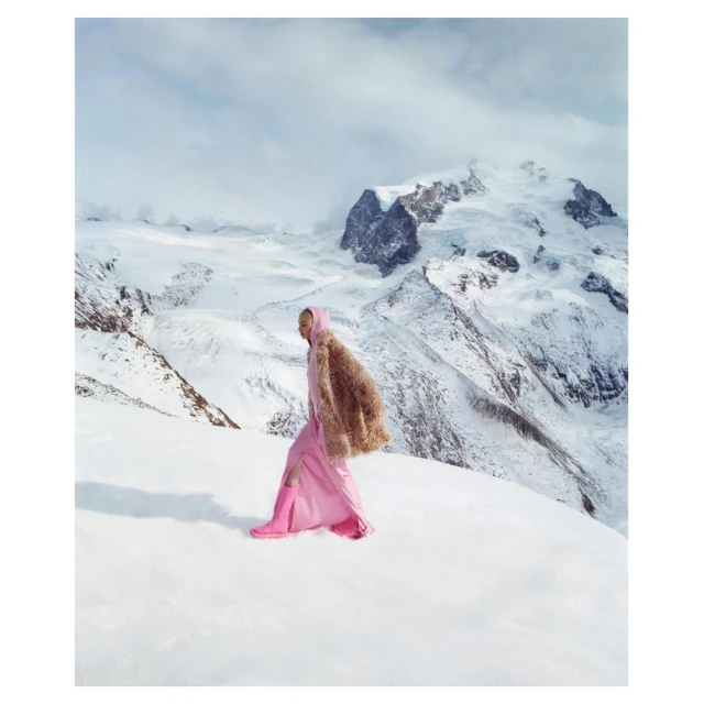 Fashionable woman in a pink dress and fur coat walks on snow-covered ground with snow-capped mountains and a bright sky in the background.