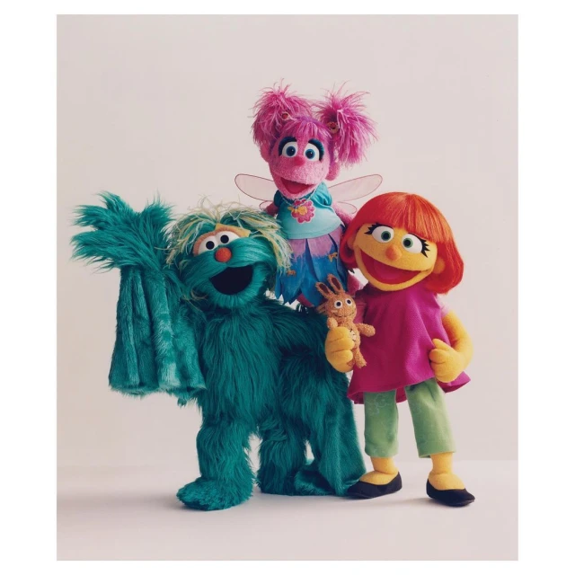 Three Sesame Street puppets, including Abby Cadabby, stand together in a studio setting against a neutral background with bright, soft lighting.